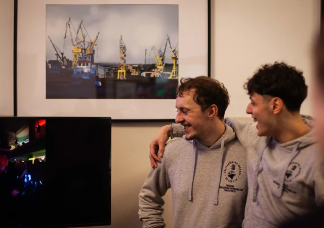 Dwóch studentów w bluzach z logo Wydziału Inżynieryjno-Ekonomicznego Transportu uśmiecha się i patrzy w stronę monitora, znajdującego się po ich lewej stronie.