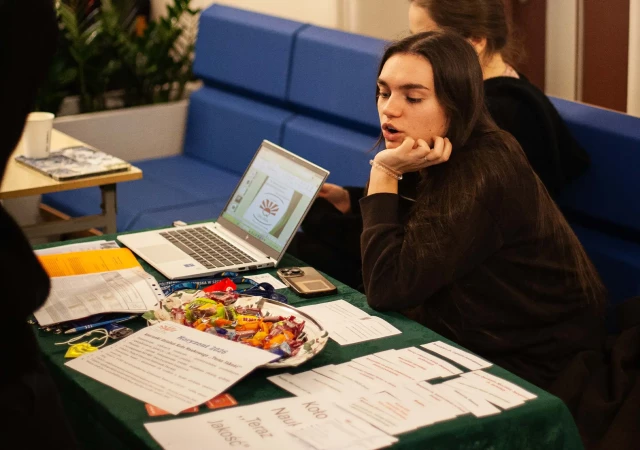 Dwie studentki z Koła Naukowego Teraz Jakość siedzą przy stole, na którym znajdują się materiały prezentujące Koło oraz laptop.