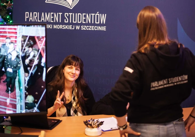 Dwie przedstawicieli Parlamentu Studentów Politechniki Morskiej: jedna studentka siedzy przy stole i uśmiecha się do aparatu, druga stoi tyłem do aparatu. W tle logo Parlamentu Studentów.