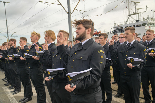 Mundurowi studenci I roku PM, na tle statku Nawigator XXI, trzymając czapki na dłoni ślubują podczas uroczystości inauguracji nowego roku akademickiego 2023/24