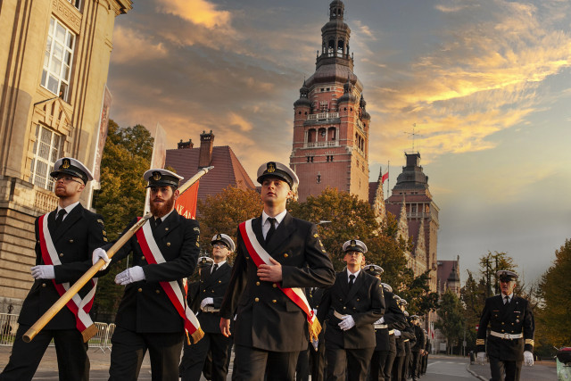 Poczet sztandarowy Politechniki Morskiej maszeruje w światle wschodzącego słońca na tle budynku Urzędu Wojewódzkiego w Szczecinie